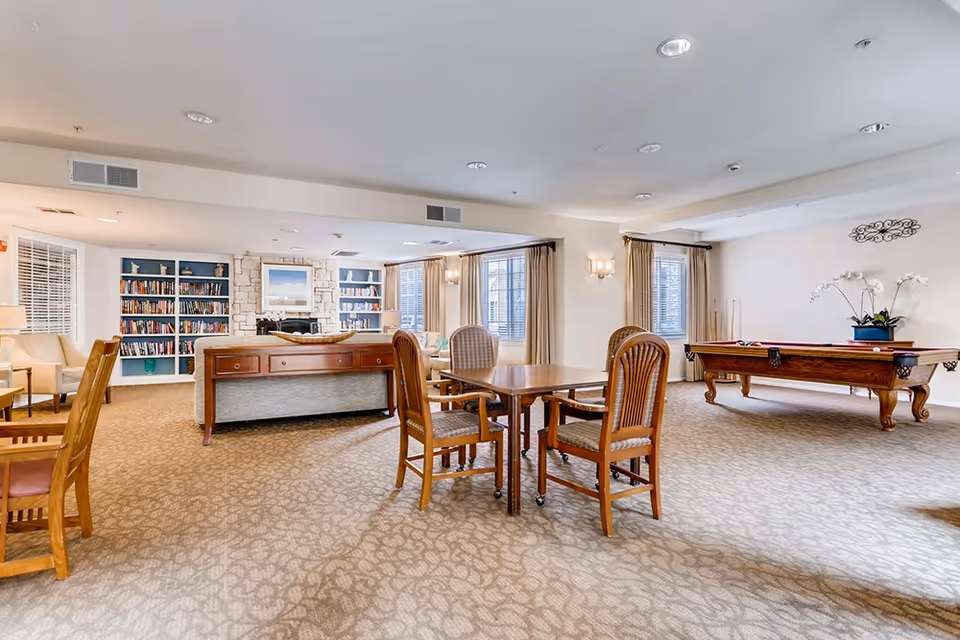 A spacious senior living common area with carpeted floors, wooden tables and chairs, a pool table, and a cozy seating area with a stone fireplace and built-in bookshelves. Large windows with beige curtains allow natural light to fill the room.