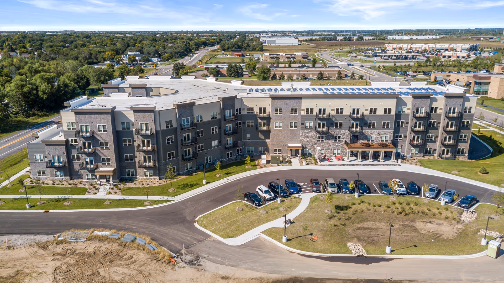 Aerial view of a four-story senior living building with balconies, a curved entrance drive, parked cars, and surrounding roads and greenery.