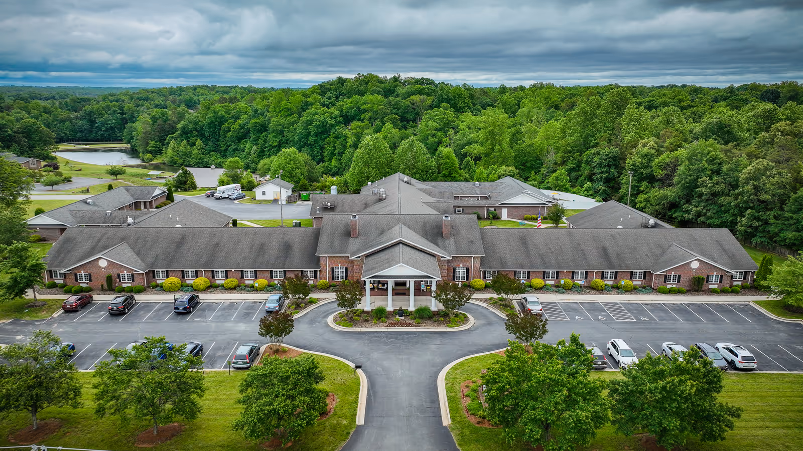 Aerial view of Mallard Ridge Assisted Living facility showing a large single-story brick building with a covered entrance, surrounded by parking lots, green lawns, trees, and a wooded area in the background under a cloudy sky.