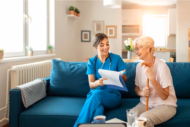 A young female healthcare professional in blue scrubs sitting on a blue couch, smiling and showing documents to an elderly woman with short white hair who is holding a wooden cane. They are in a bright, cozy living room with a kitchen visible in the background.