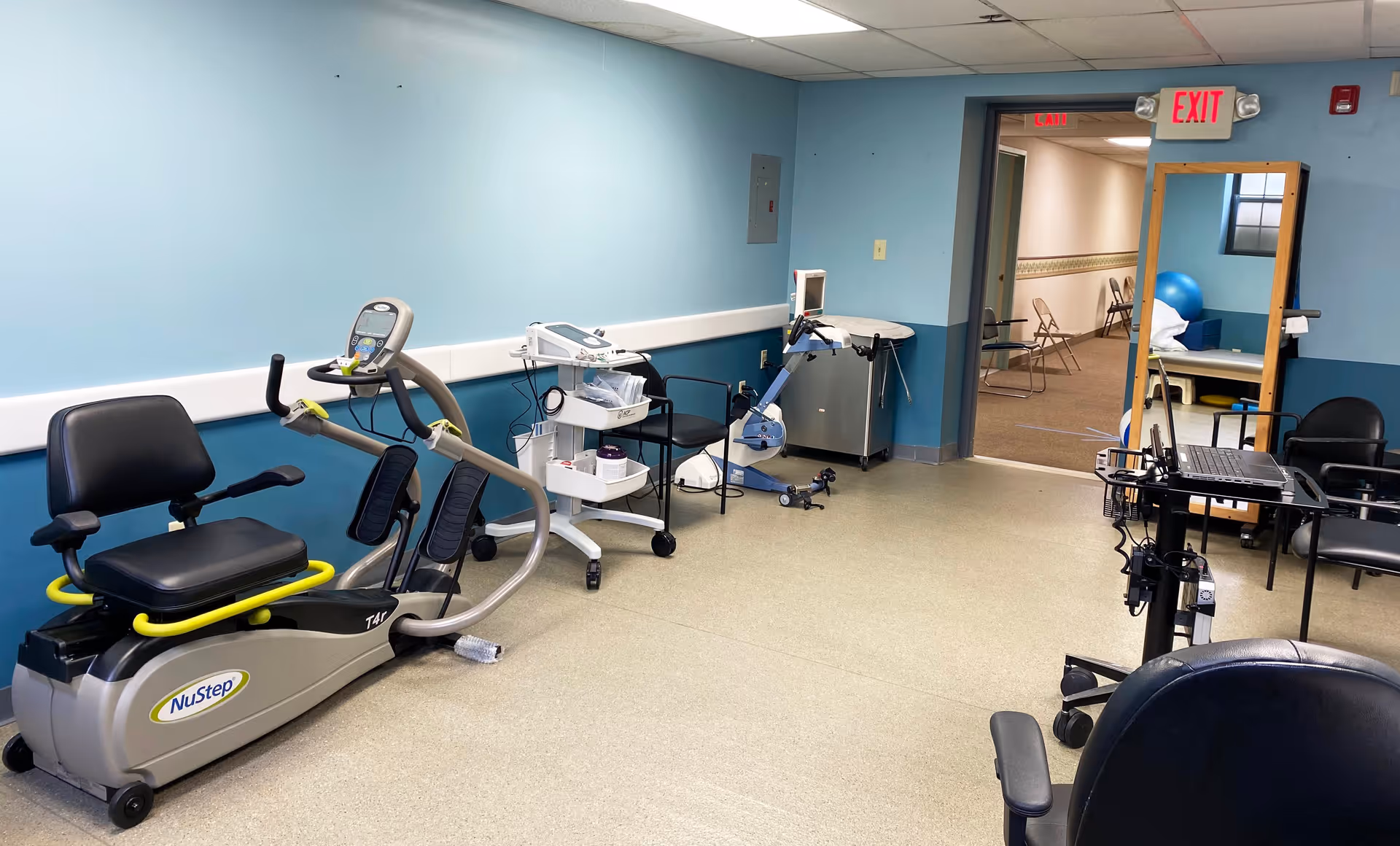 A rehabilitation or physical therapy room with exercise equipment including a NuStep recumbent cross trainer, a stationary arm bike, chairs, a rolling cart with medical supplies, and a large mirror. The room has blue walls and a beige floor, with an exit sign above a doorway leading to another room with chairs and a blue exercise ball.
