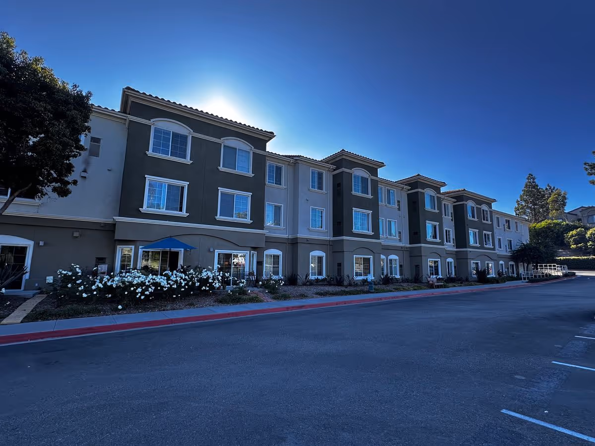 Three-story residential building front with landscaped flowerbeds, a parking area, and a clear blue sky.