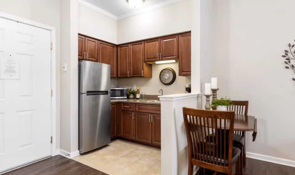 A compact kitchen area with wooden cabinets, a stainless steel refrigerator, a microwave, and a small sink. Adjacent to the kitchen is a small dining table with two wooden chairs, decorated with candles and a small plant. The walls are painted light beige, and the floor transitions from tile in the kitchen to wood in the dining area.