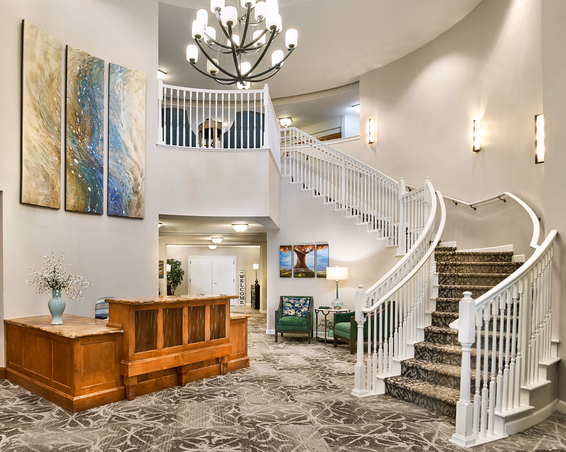 Interior view of a senior living facility lobby featuring a curved staircase with white railings and carpeted steps. There is a wooden reception desk with a vase of flowers on it, patterned carpet flooring, and modern wall sconces providing lighting. The space includes a seating area with green chairs, a side table with a lamp, and decorative paintings on the walls. A chandelier hangs from the ceiling above the staircase.