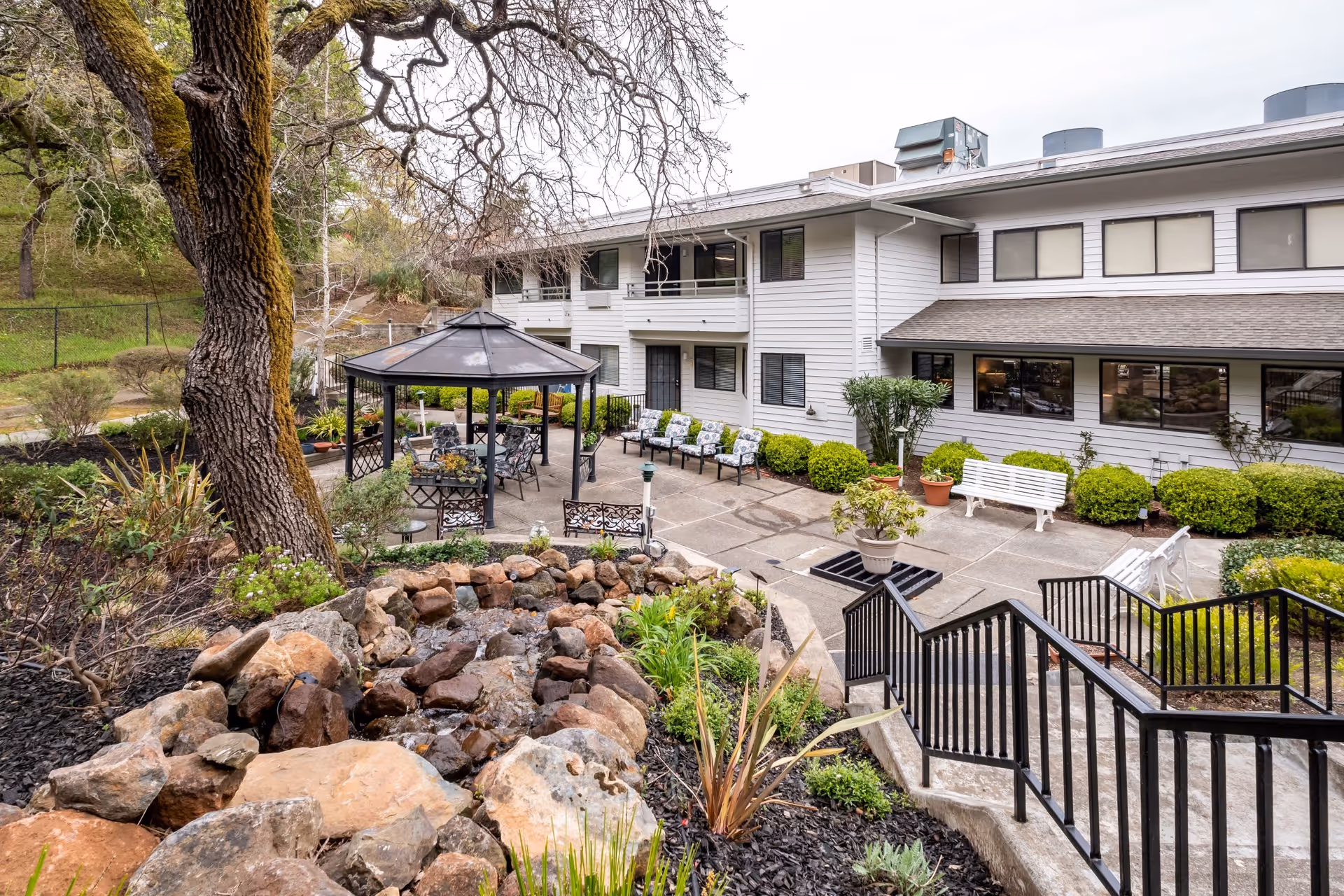 Outdoor patio area at Deer Park Retirement Community featuring a gazebo with seating, multiple benches, potted plants, a stone-lined water feature, and a two-story building in the background surrounded by greenery.
