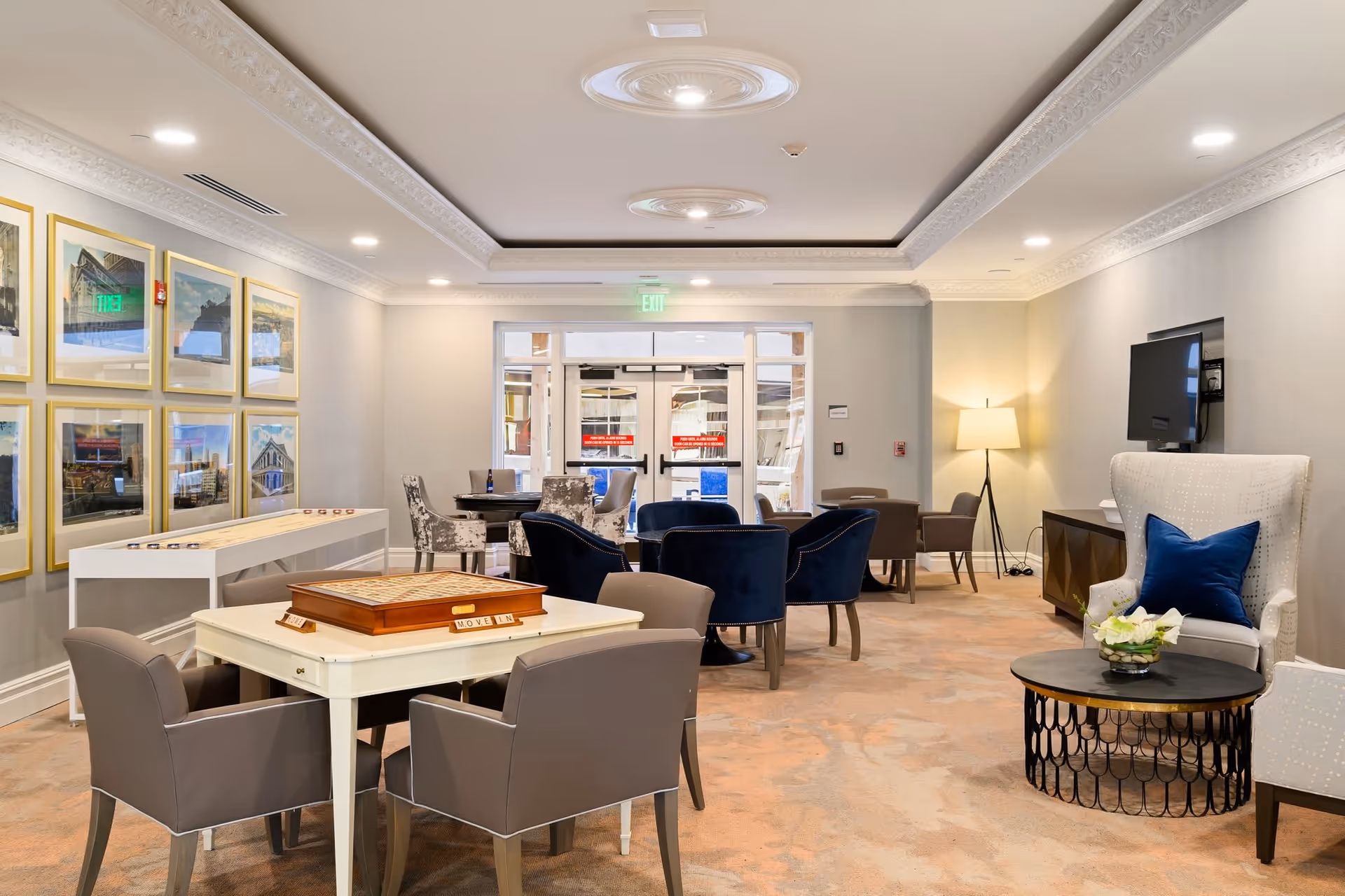 A well-lit lounge area with multiple seating arrangements including gray and blue chairs around tables. A shuffleboard table is positioned against the left wall, which is decorated with framed pictures. A TV is mounted on the right wall above a cabinet, and a floor lamp provides additional lighting. The room has a beige carpet and a decorative ceiling with recessed lighting.