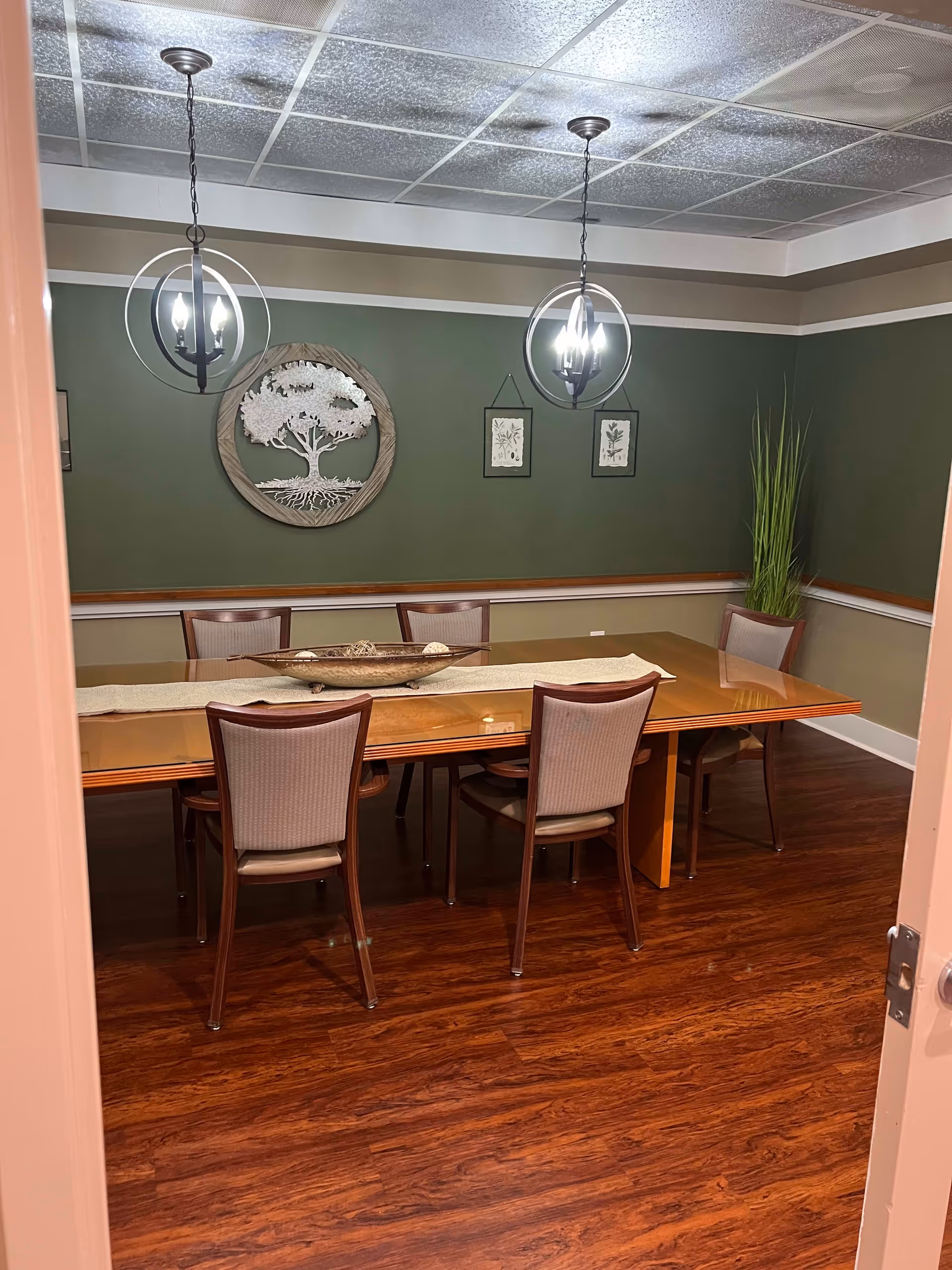 A dining room with a wooden table and six chairs. The walls are painted green with white trim, and there are two modern pendant lights hanging from the ceiling. On the wall, there is a decorative wooden tree art piece and two framed botanical prints. A tall plant is placed in the corner of the room.
