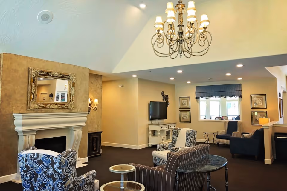 A cozy living room area with a white fireplace topped by a decorative gold-framed mirror. The room features patterned and striped armchairs, a small round glass-top table, a flat-screen TV on a white stand, and a large chandelier hanging from a vaulted ceiling. The walls are painted beige, and there is a window with a valance letting in natural light.