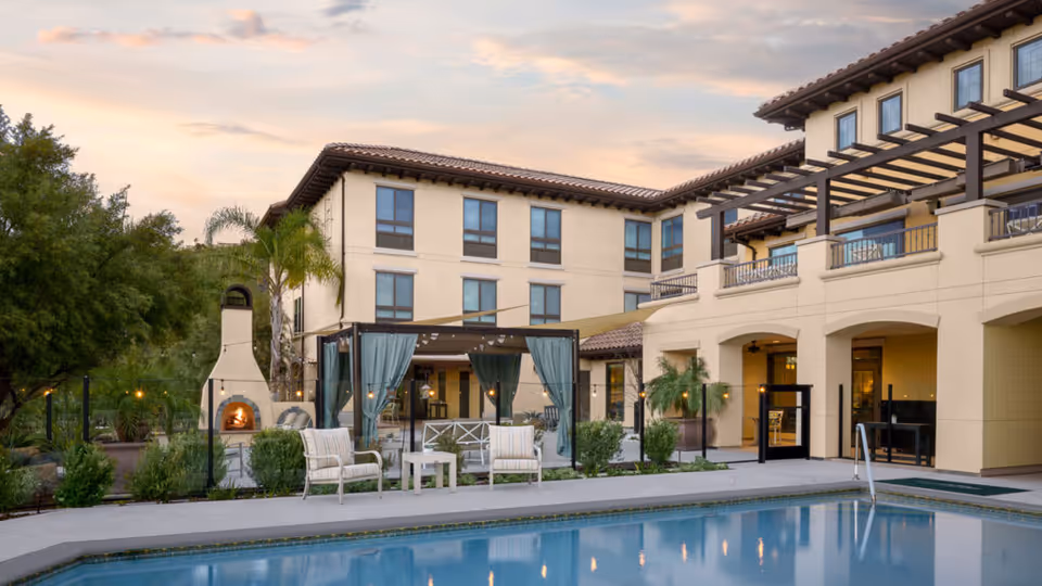 Outdoor area of Belmont Village Senior Living Calabasas featuring a swimming pool in the foreground, lounge chairs, a pergola with curtains, a fireplace, and a multi-story building with balconies in the background under a partly cloudy sky at sunset.