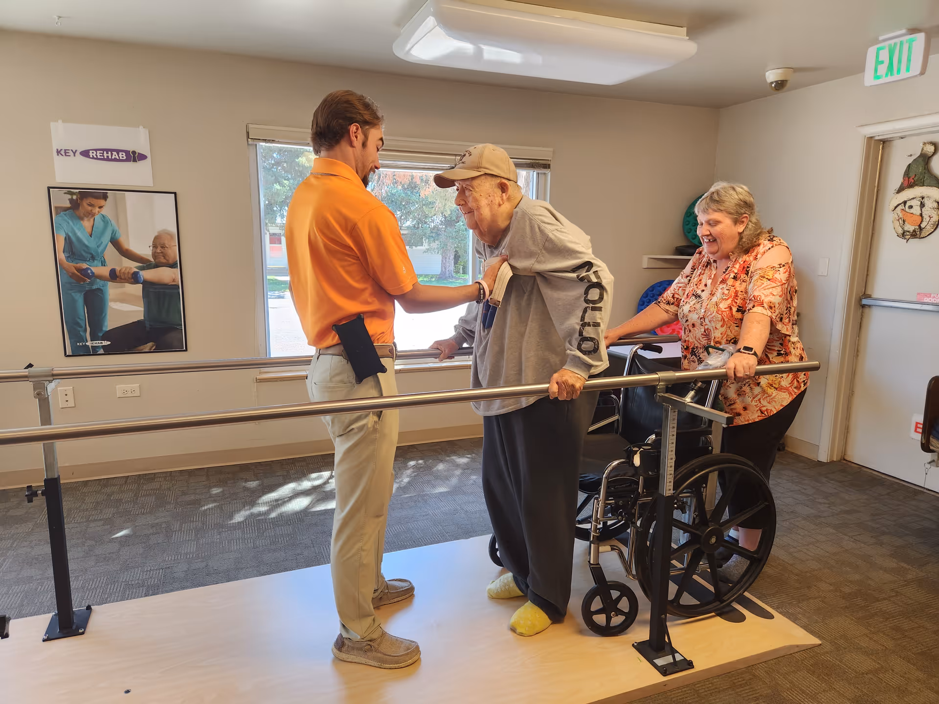 An elderly man wearing a cap and gray sweatshirt is supported by a man in an orange shirt as he practices walking between parallel bars in a rehabilitation room. A woman stands behind the elderly man, smiling and holding a wheelchair. The room has a window, a poster on the wall, and an exit sign above a door.