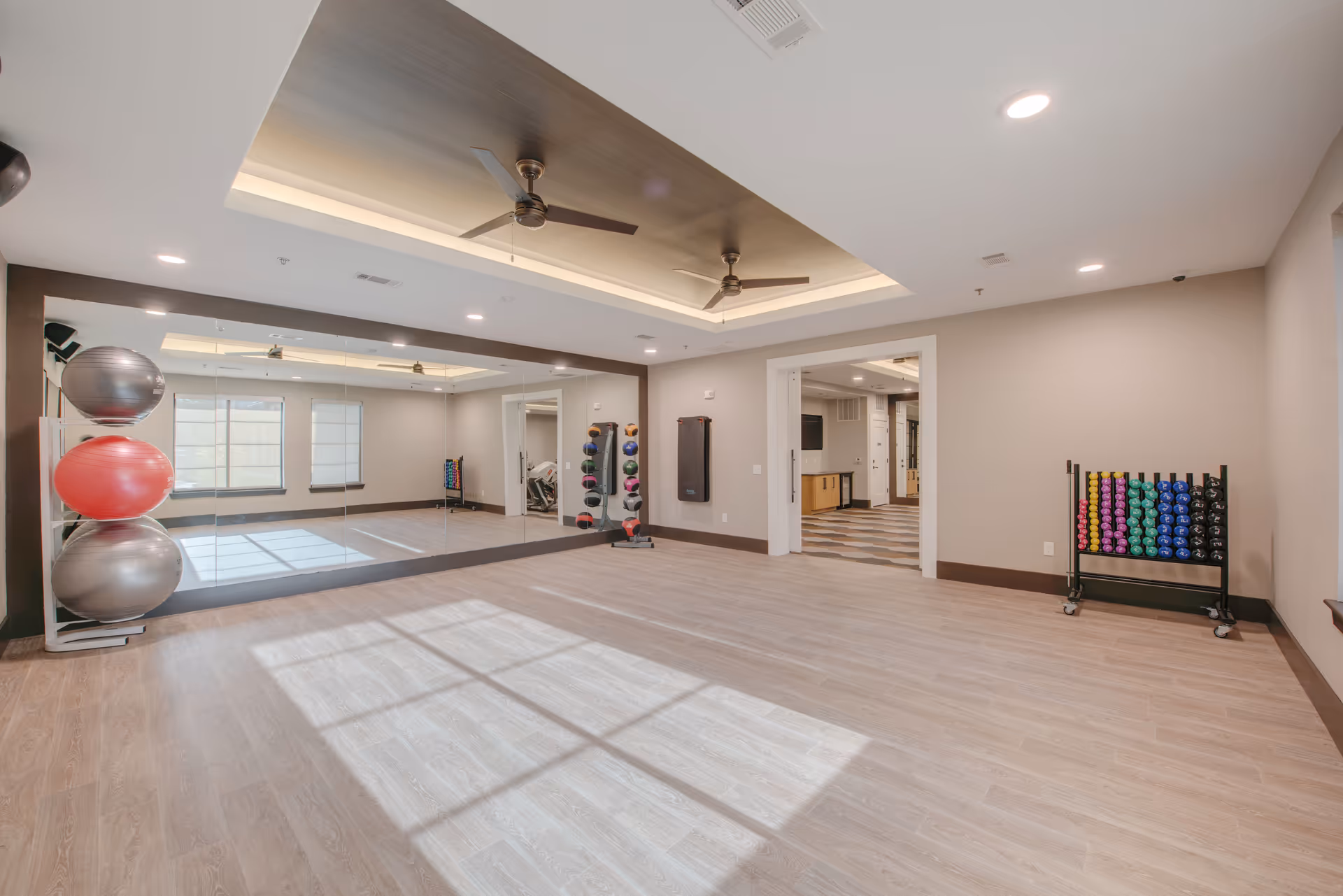 A spacious exercise room with light wood flooring, large wall mirrors, and ceiling fans. The room contains racks holding colorful exercise balls and weights. Natural light streams in through windows, and an open doorway leads to another room with a checkered floor.