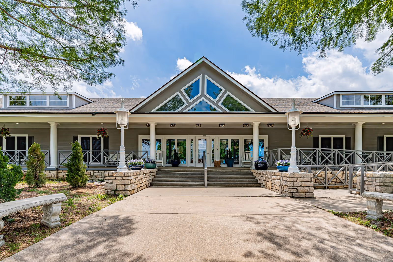 Front exterior view of The Homestead of Denison building with a wide entrance featuring steps, columns, large windows, and a triangular window design at the roof peak. There are stone planters with flowers, hanging flower baskets, and benches on either side of the walkway leading to the entrance. Trees frame the scene under a partly cloudy blue sky.