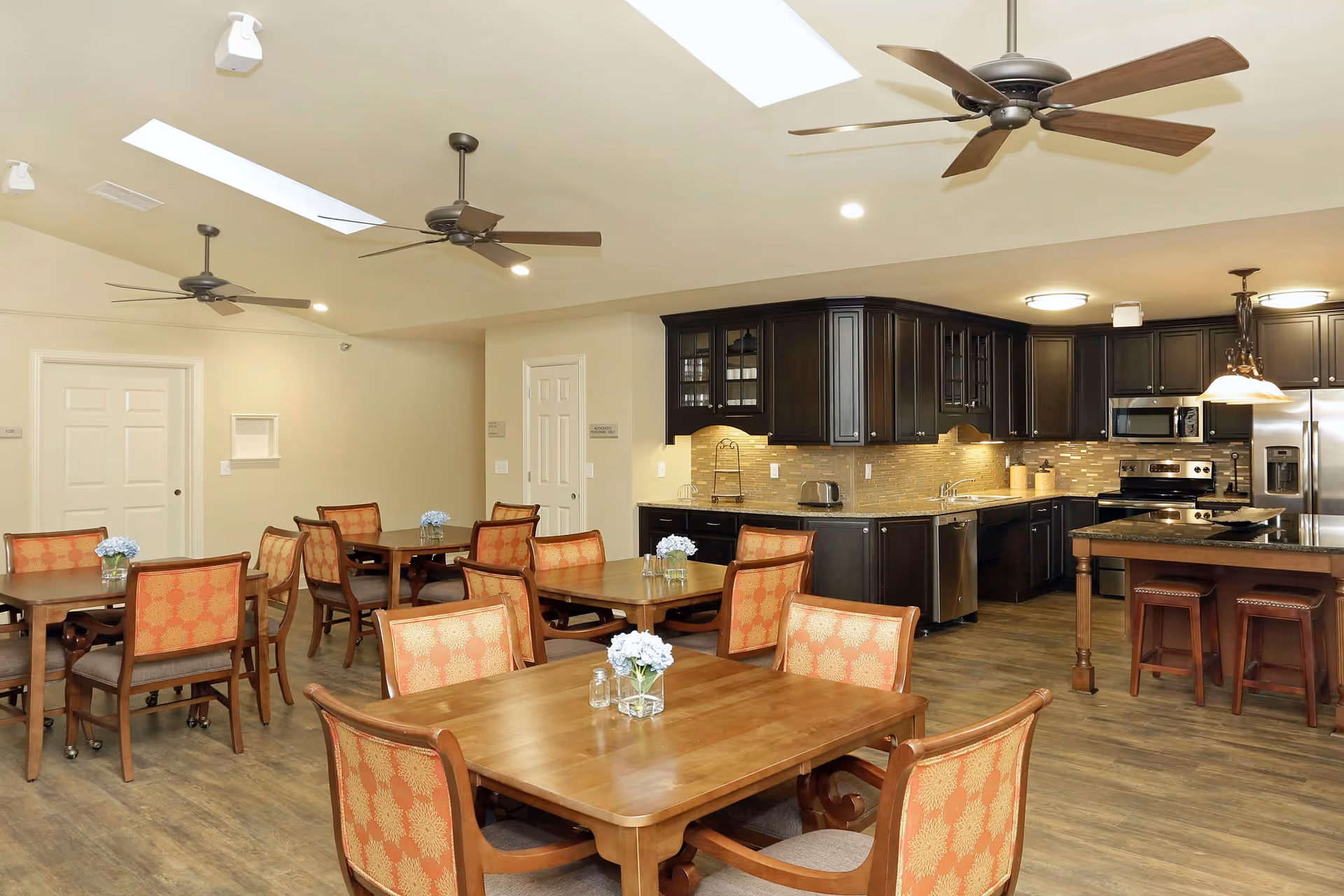 A spacious dining area with several wooden tables and chairs featuring orange patterned upholstery. The room has a modern kitchen with dark wood cabinets, stainless steel appliances, and a kitchen island with stools. The ceiling has recessed lighting, ceiling fans, and skylights, creating a bright and welcoming atmosphere.