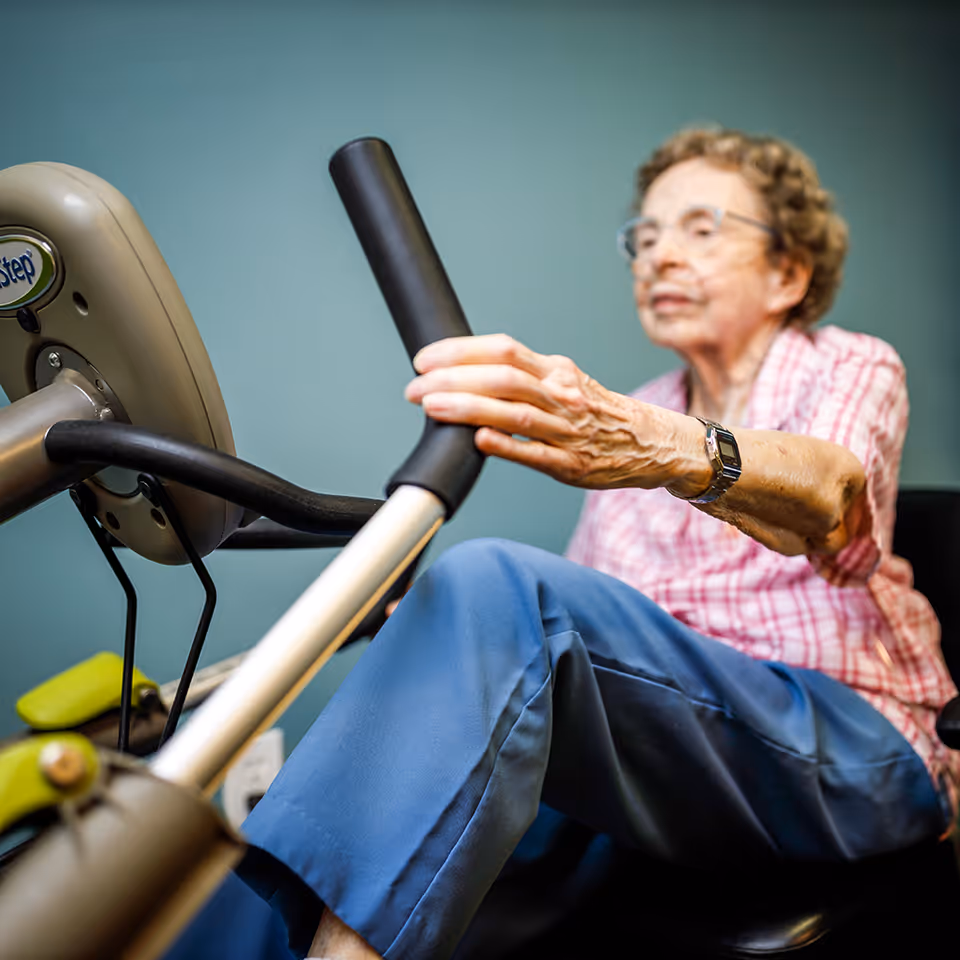 An elderly woman wearing glasses and a pink checkered shirt is using a seated exercise machine, gripping the handles and pedaling with her legs in a fitness or rehabilitation setting.