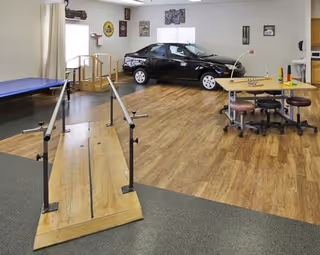Rehabilitation therapy room with a wooden parallel-bars ramp, a black practice car, therapy table and stools, and exercise mat on wood flooring.