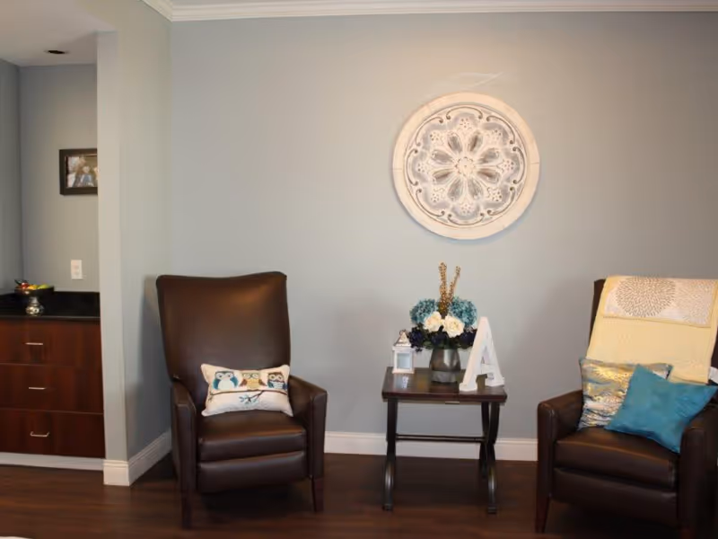 A cozy sitting area with two brown leather armchairs separated by a small wooden side table. The table holds a flower arrangement, a decorative letter 'A', and a small lantern. A round decorative wall piece hangs on the light gray wall above the table. The floor is dark wood, and there is a small cabinet with drawers visible to the left.