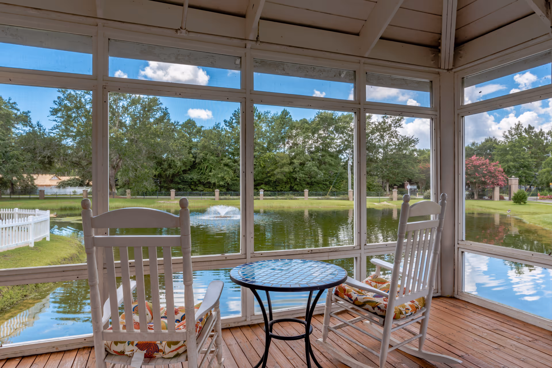 A screened-in porch with two white rocking chairs with floral cushions and a small round table between them, overlooking a pond with a fountain and surrounded by green trees and a white picket fence under a blue sky with scattered clouds.