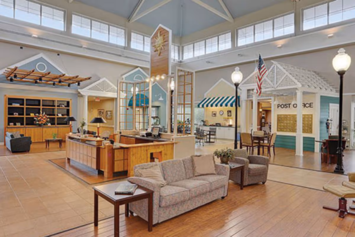 Spacious, brightly lit senior living facility common area with seating, a reception desk, and a mock post office storefront.