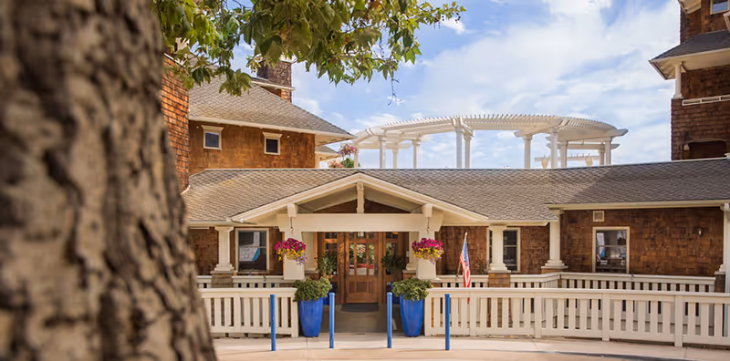 Front exterior view of Crown Cove facility showing a brown shingle-style building with a covered entrance, white railing, blue planters with flowers, and an American flag. A tree trunk is visible in the foreground on the left side.
