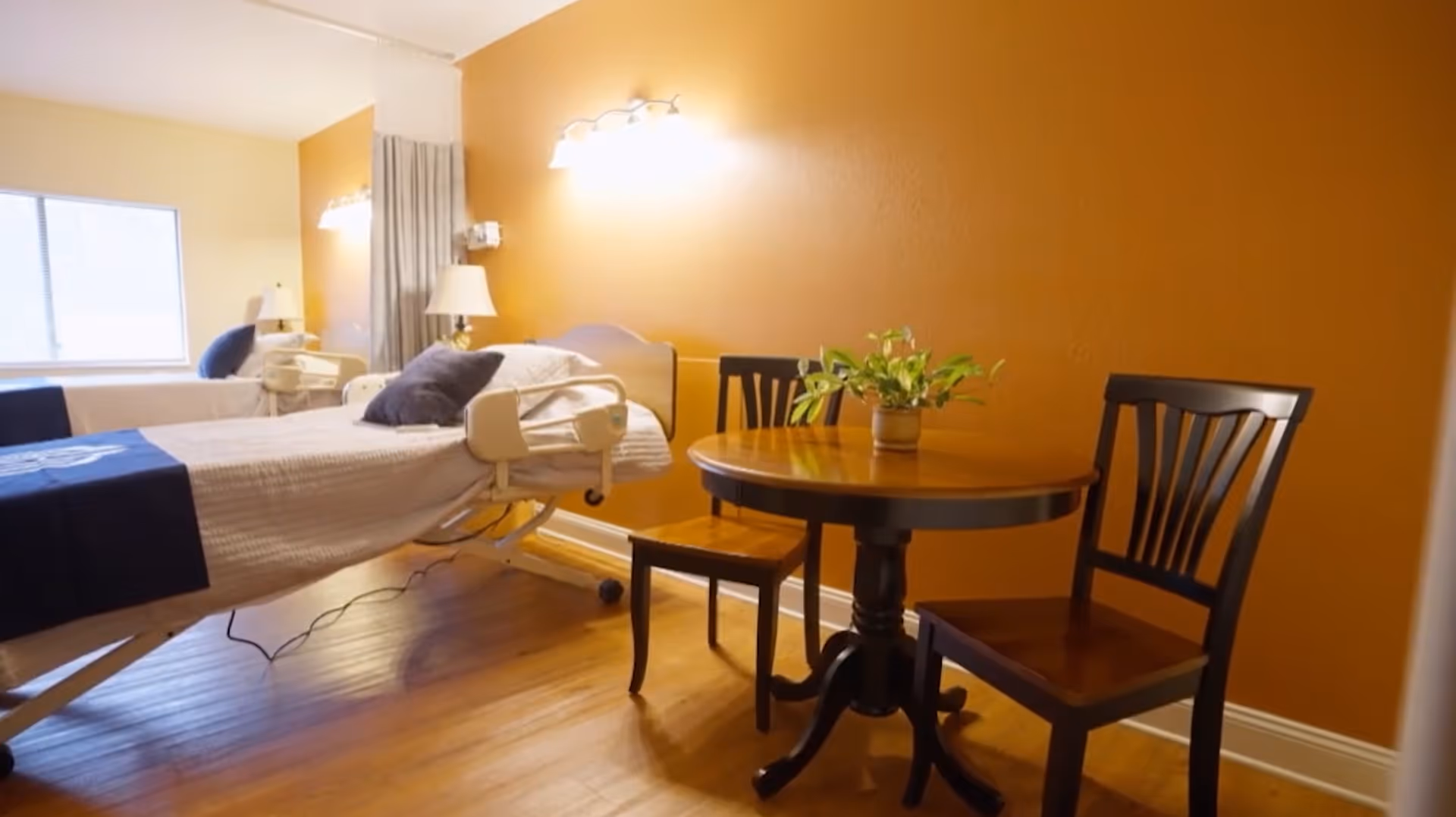 A room with two hospital-style beds, each with white bedding and dark blue blankets. There is a small round wooden table with two wooden chairs next to an orange wall. A small potted plant is on the table. The room has wooden flooring and a window letting in natural light.