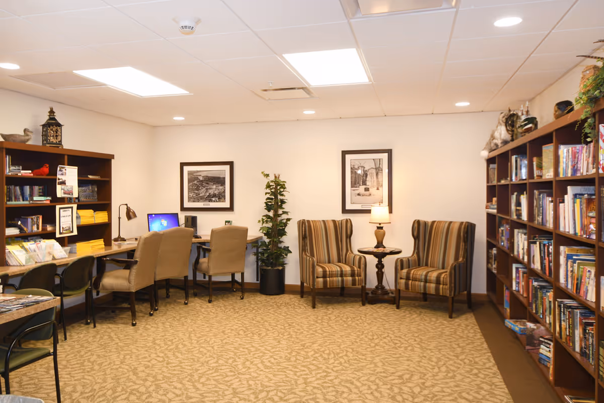 A cozy library or reading room with bookshelves filled with books on the right and left sides. There are two striped armchairs with a small round table and lamp between them against the back wall. On the left side, there are desks with chairs and a computer, along with framed black and white pictures on the wall and a potted plant.
