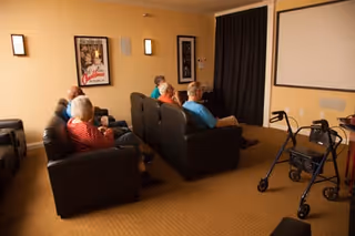 A group of elderly people seated on black leather couches in a small theater room watching a movie projected on a white screen. There are framed pictures on the beige walls and a walker is positioned near the seating area.