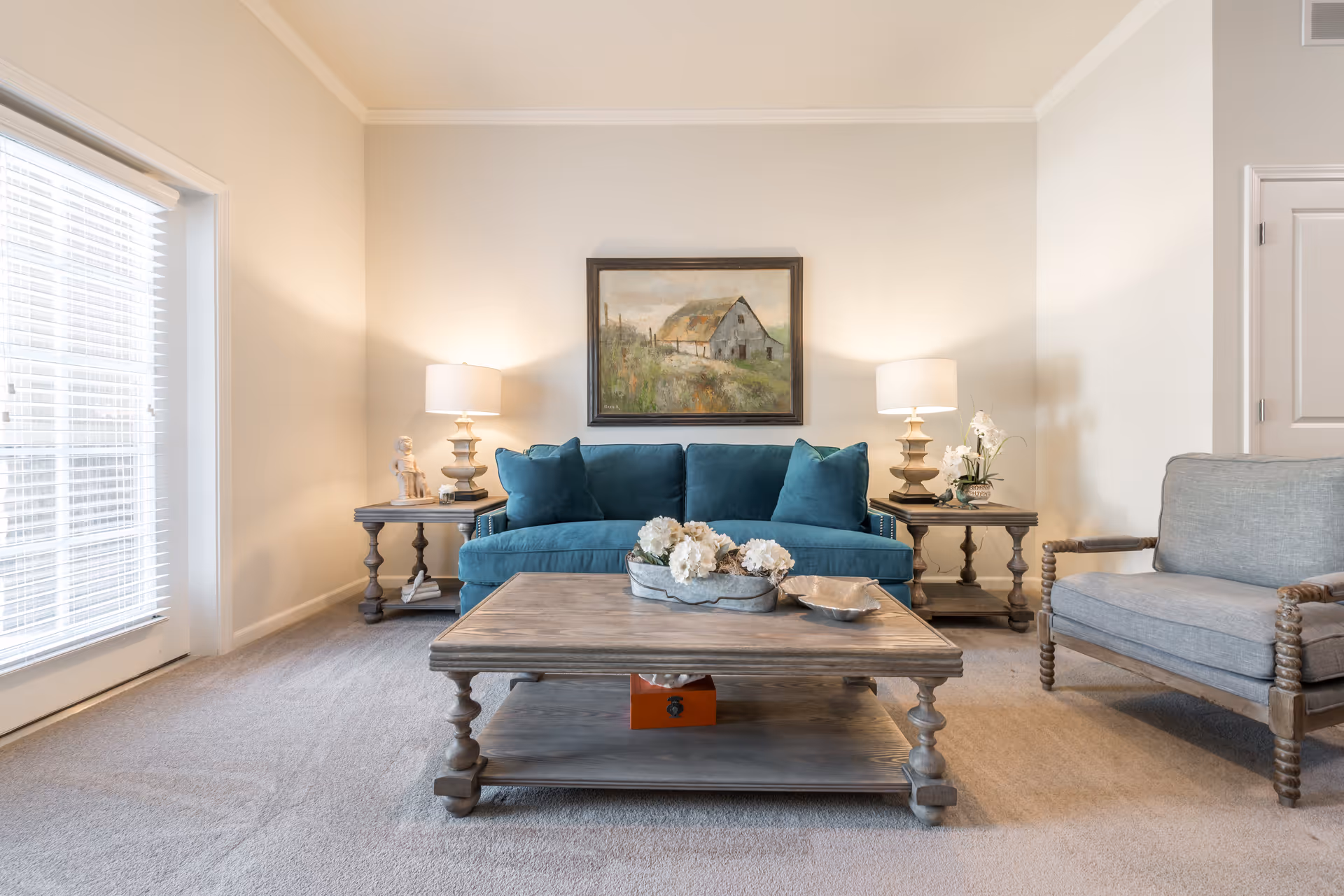 A cozy living room with a teal sofa centered against a beige wall, flanked by two matching wooden side tables each with a lamp. A wooden coffee table with decorative flowers sits in front of the sofa. To the right, there is a cushioned armchair with wooden armrests. A large window with blinds is on the left side, and a framed painting of a barn hangs above the sofa.