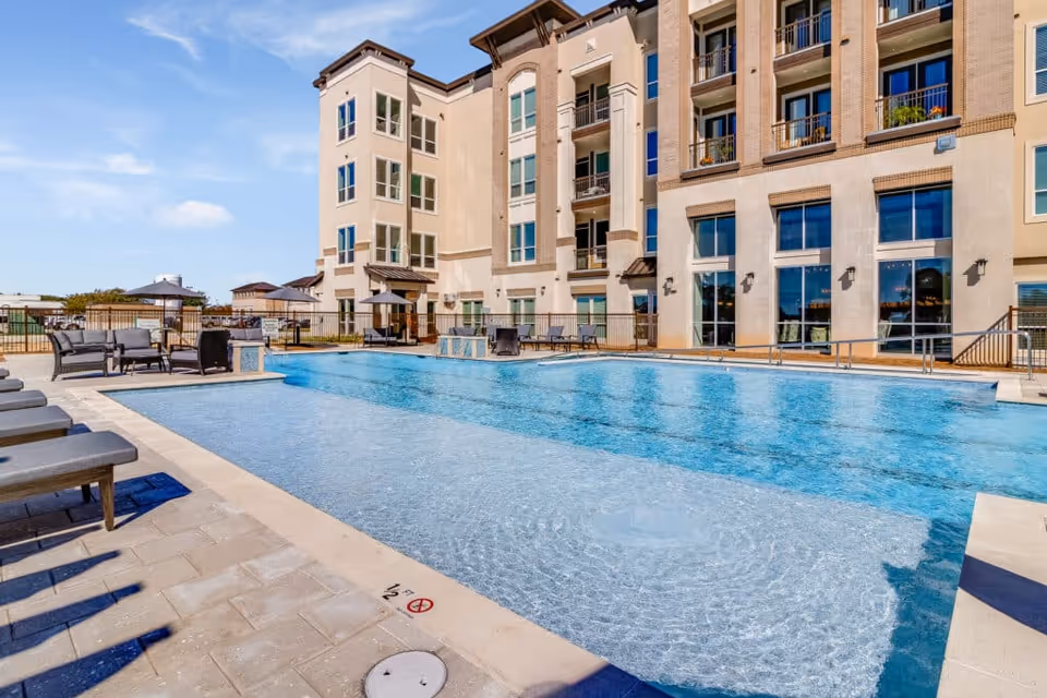 Outdoor swimming pool with lounge chairs and seating area beside a multi-story senior living building.