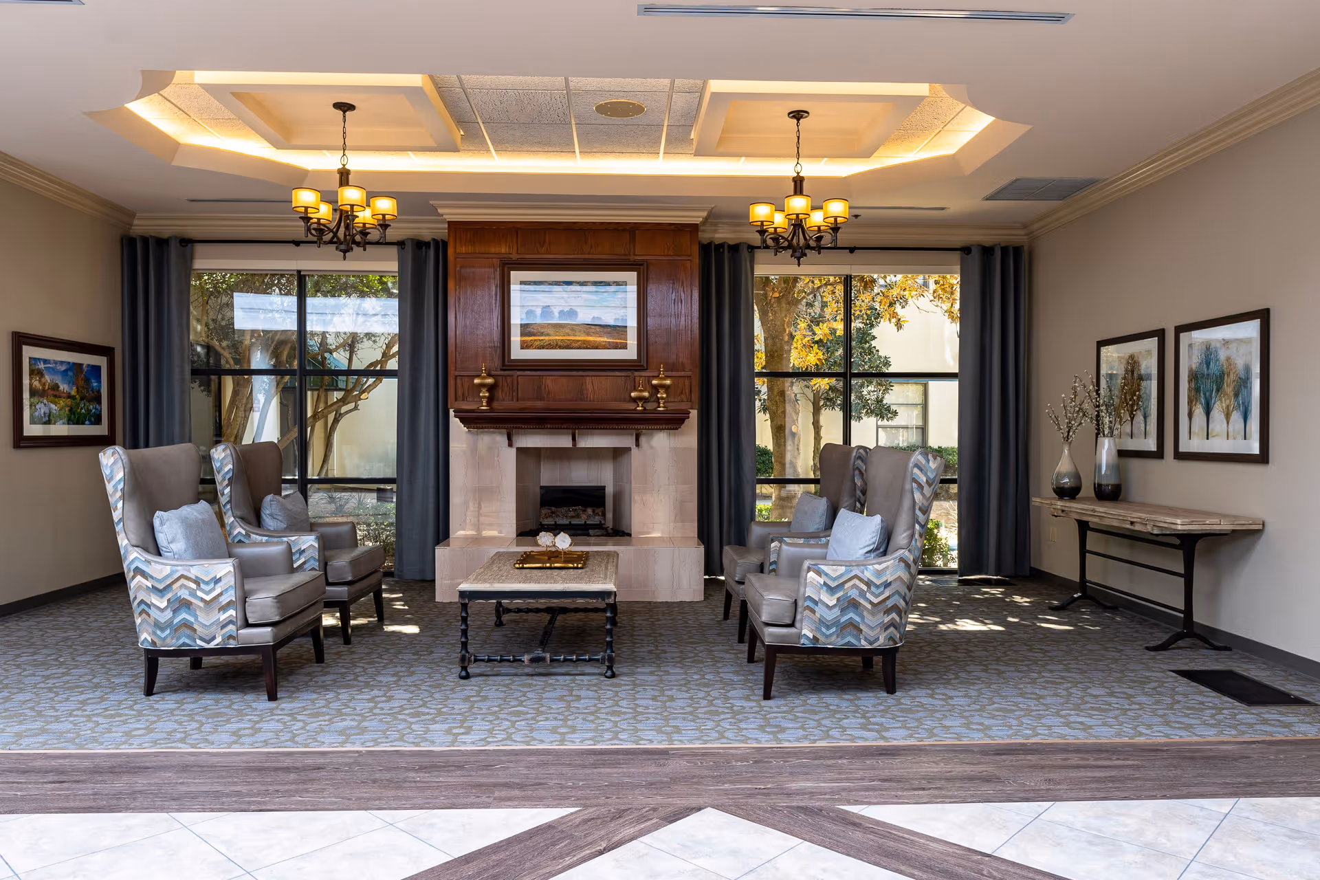A cozy and elegant common area in a senior living facility featuring a central fireplace with a wooden mantel, flanked by large windows with dark curtains. There are four patterned armchairs arranged around a small coffee table on a carpeted floor. The room is decorated with framed artwork on the walls and a console table with vases and decorative branches. Two chandeliers hang from a recessed ceiling with soft lighting.