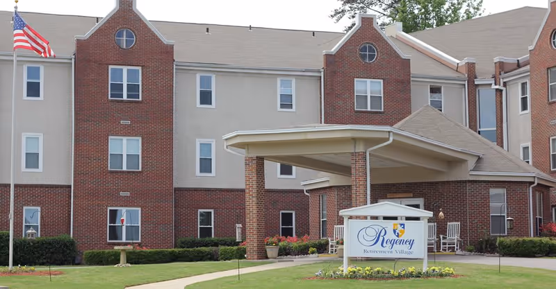 Exterior view of Regency Retirement Village of Birmingham, showing a large brick and beige building with multiple windows, a covered entrance supported by brick columns, an American flag on a flagpole, and a sign with the facility name in front surrounded by a small garden.