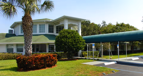Exterior view of a two-story building with a green roof and white walls, surrounded by palm trees, bushes, and a well-maintained lawn. There is a covered walkway leading to the entrance and a parking area with handicap parking spaces in front.