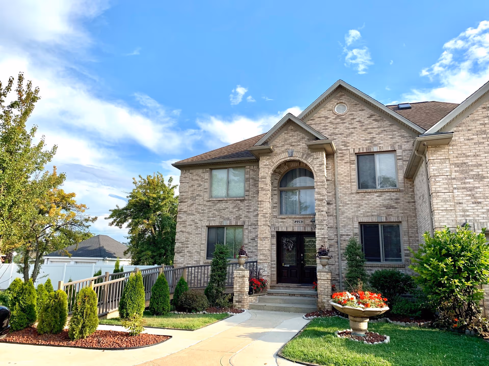 Front exterior view of a two-story brick building with a dark brown front door, arched window above the entrance, and several windows. The building is surrounded by a well-maintained lawn, shrubs, small trees, and a flower planter with red and white flowers. A concrete walkway leads to the entrance, and a blue sky with some clouds is visible above.