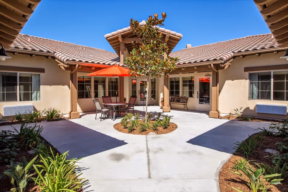 Outdoor courtyard area of a senior living facility with a central tree surrounded by plants, concrete pathways, and a seating area with a table, chairs, and a red umbrella. The building has beige walls and a tiled roof under a clear blue sky.
