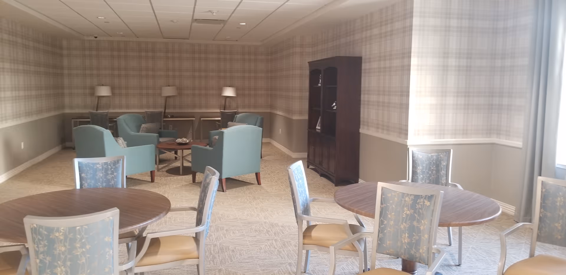 A well-lit room with round wooden tables and chairs with floral upholstery in the foreground, and a seating area with blue armchairs and floor lamps in the background. The walls have a plaid wallpaper design and there is a dark wooden cabinet against one wall.