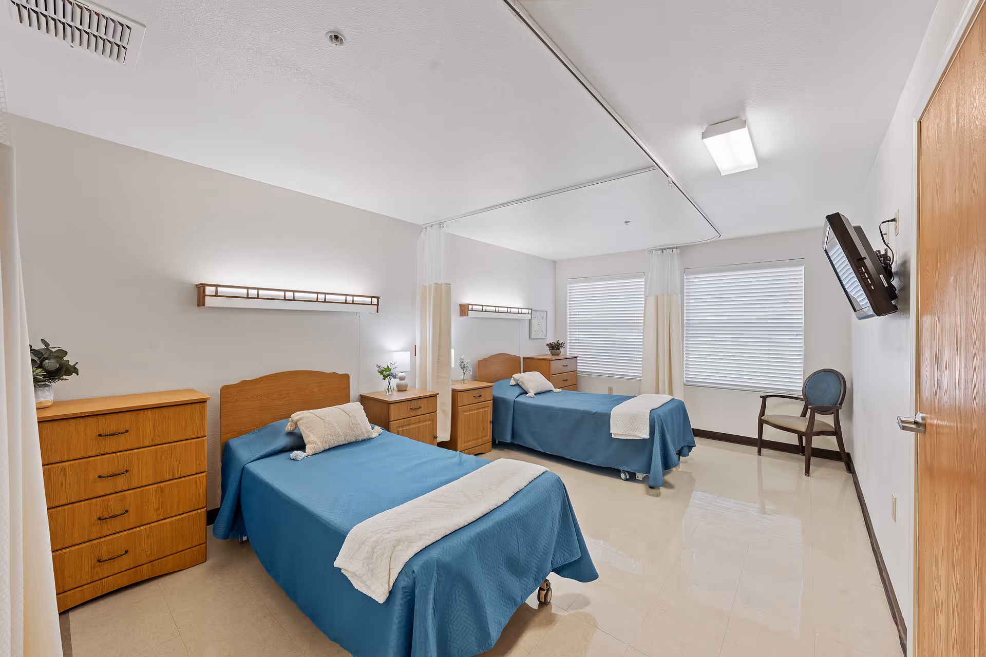 A clean and bright room with two single beds covered in blue bedspreads and white blankets. Each bed has a wooden headboard and a small wooden nightstand with a lamp and decorative items. There is a wooden dresser on the left side and a chair near two large windows with blinds. A flat-screen TV is mounted on the wall opposite the beds. The room has a tiled floor and white walls with ceiling lights.