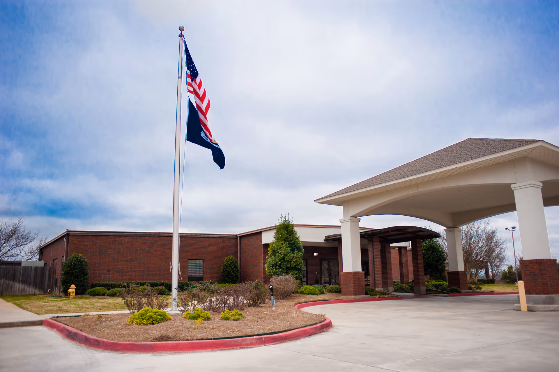 Exterior view of Riverview Care Center showing a brick building with a covered entrance driveway, a flagpole with the American flag and another flag, some bushes, and a cloudy sky.
