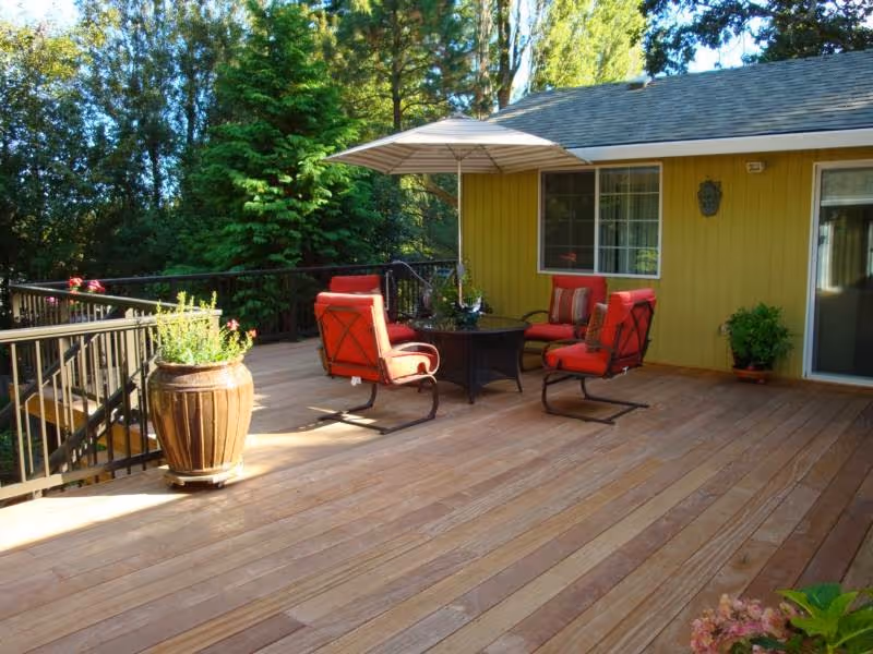 A spacious wooden deck attached to a yellow house, featuring four red cushioned chairs around a round table with a large umbrella. There are potted plants on the deck and trees in the background.