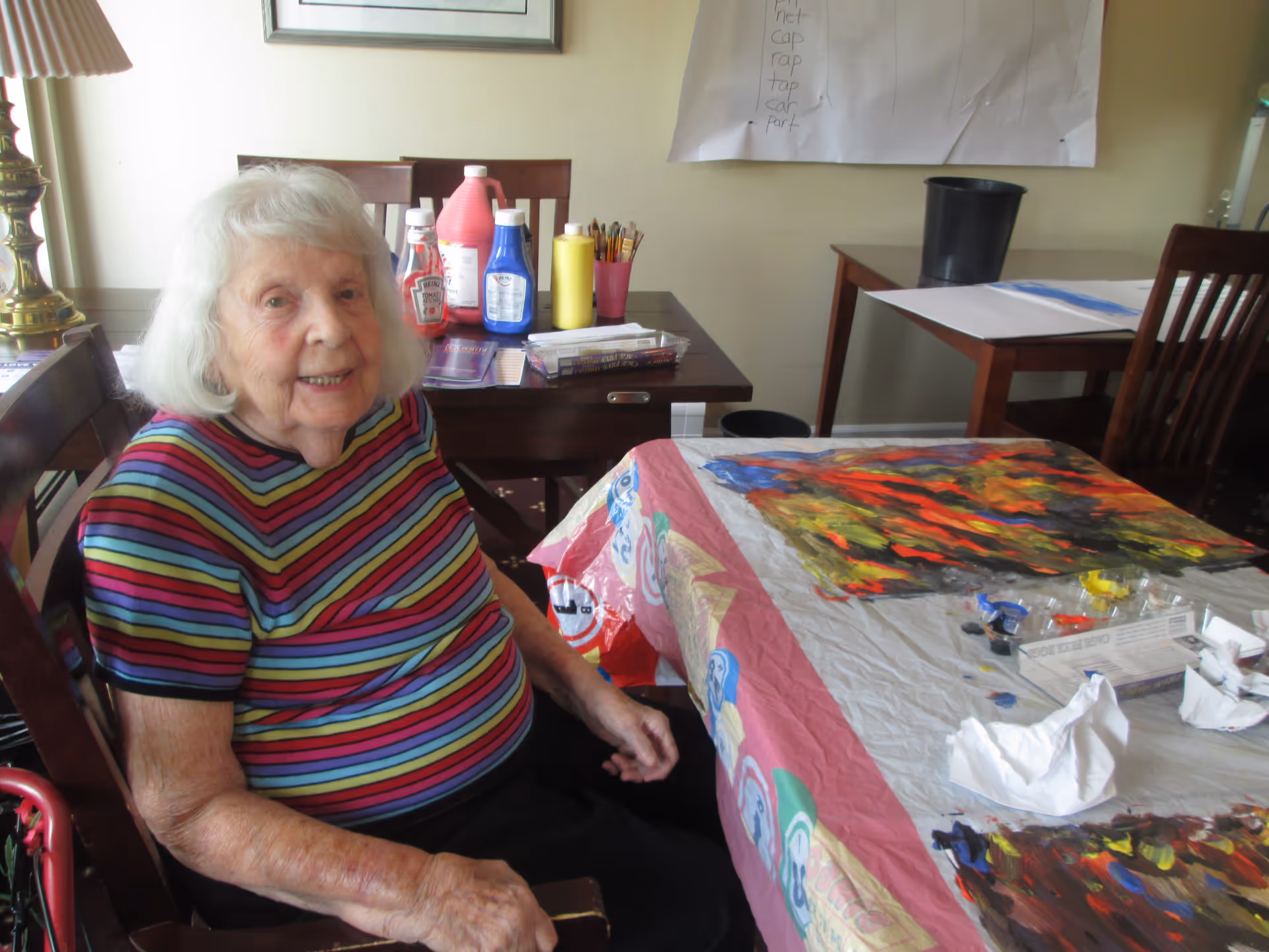 An elderly woman with white hair wearing a colorful striped shirt sits at a table covered with a plastic tablecloth and art supplies, including paint and brushes. Behind her are two wooden chairs and a table with bottles of paint and other art materials. A large sheet of paper with handwritten words is pinned to the wall.