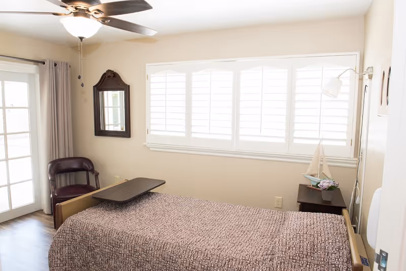 A bright and clean bedroom in a skilled nursing facility featuring a single bed with a patterned bedspread, a small overbed table, a wooden chair, a wall-mounted mirror, a ceiling fan with light, and a window with white shutters. There is also a small bedside table with a decorative sailboat and a potted plant.
