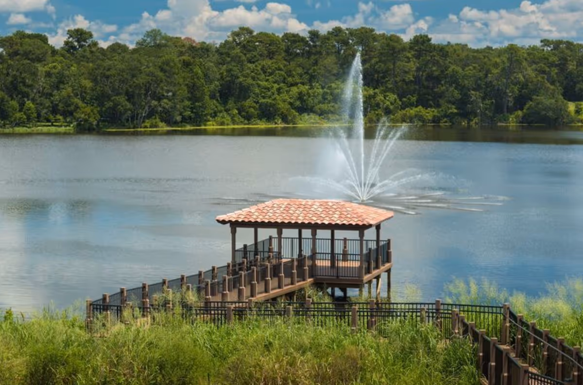 A wooden gazebo with a tiled roof on a pier extending over a lake, with a large water fountain spraying water in the background and lush green trees surrounding the lake under a partly cloudy sky.