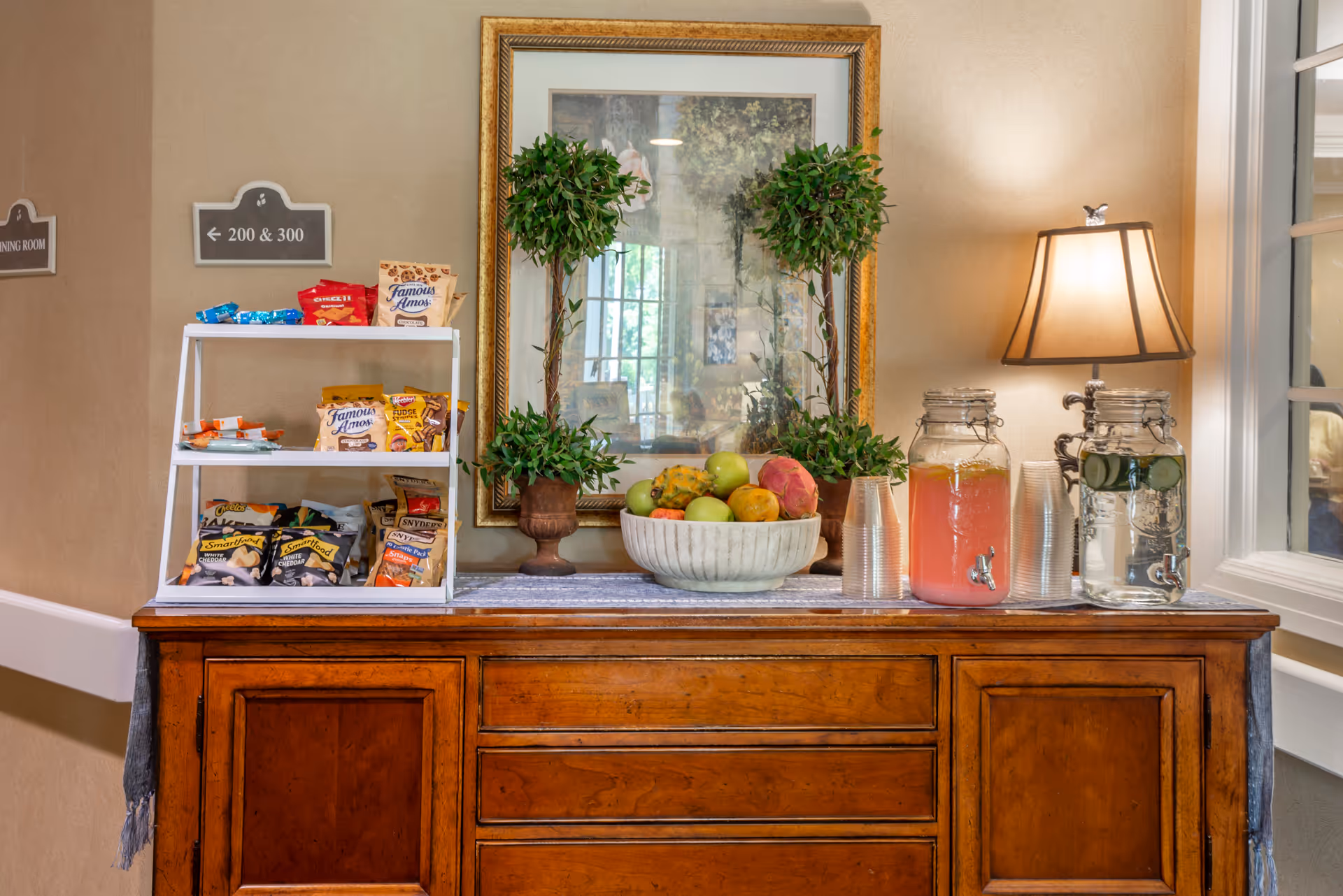 Wooden sideboard topped with a bowl of fruit, a snack display, beverage dispensers, a lamp, and decorative plants in front of a mirror.
