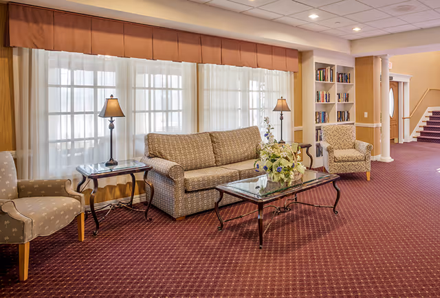Well-lit lobby seating area with a sofa, armchairs, glass coffee table with flowers, lamps, bookshelf and a staircase in the background.
