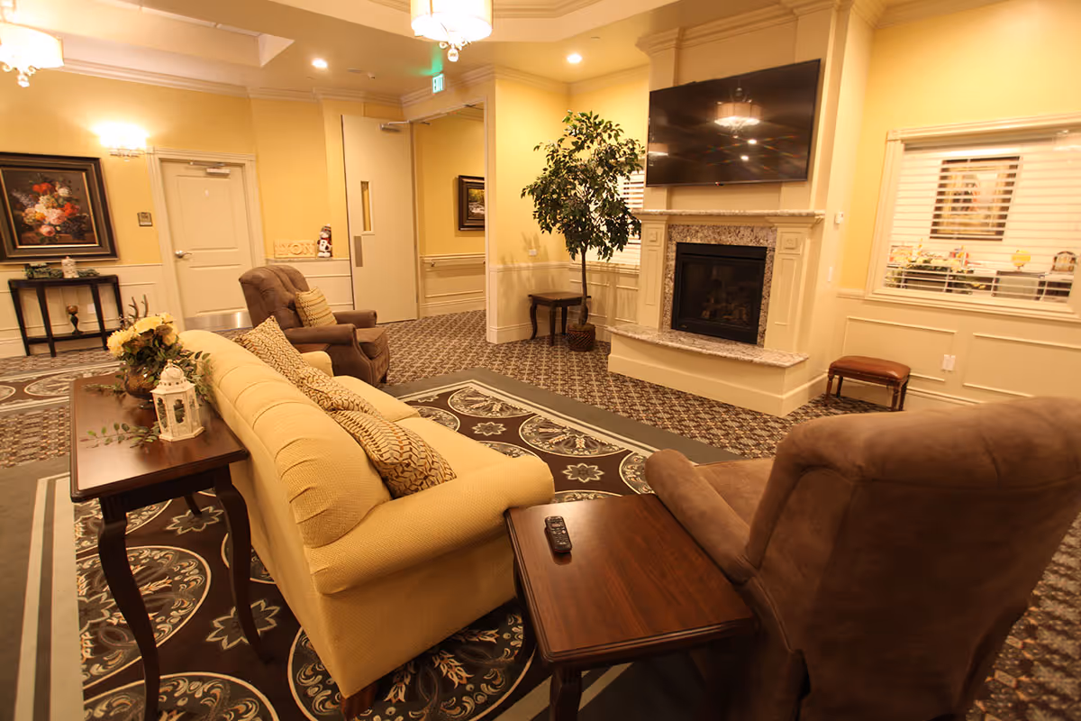 A cozy assisted living facility common area with a beige sofa, two brown armchairs, a wooden side table with a remote control, a fireplace with a mounted flat-screen TV above it, a potted plant, and framed artwork on the walls. The room has warm lighting and patterned carpet.