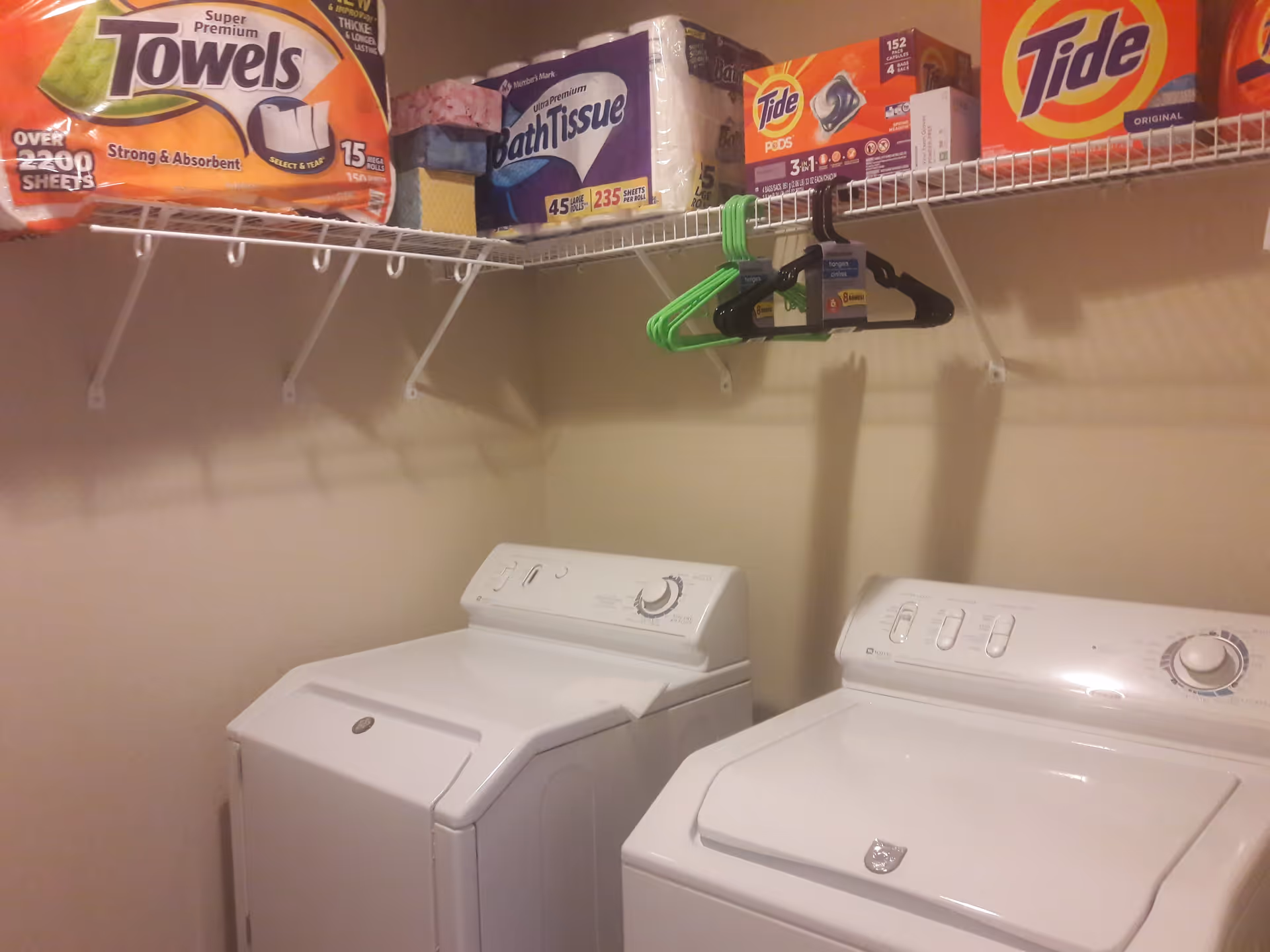 Laundry room with a white washing machine and dryer side by side. Above them is a wire shelf holding paper towels, bath tissue, laundry detergent, and several hangers.