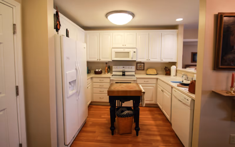 A kitchen with white cabinets and appliances, including a refrigerator, microwave, stove, and dishwasher. There is a wooden island with a basket underneath in the center of the room. The floor is wooden, and the walls are light-colored. A framed painting and a candle holder with a red candle are visible on the right side.