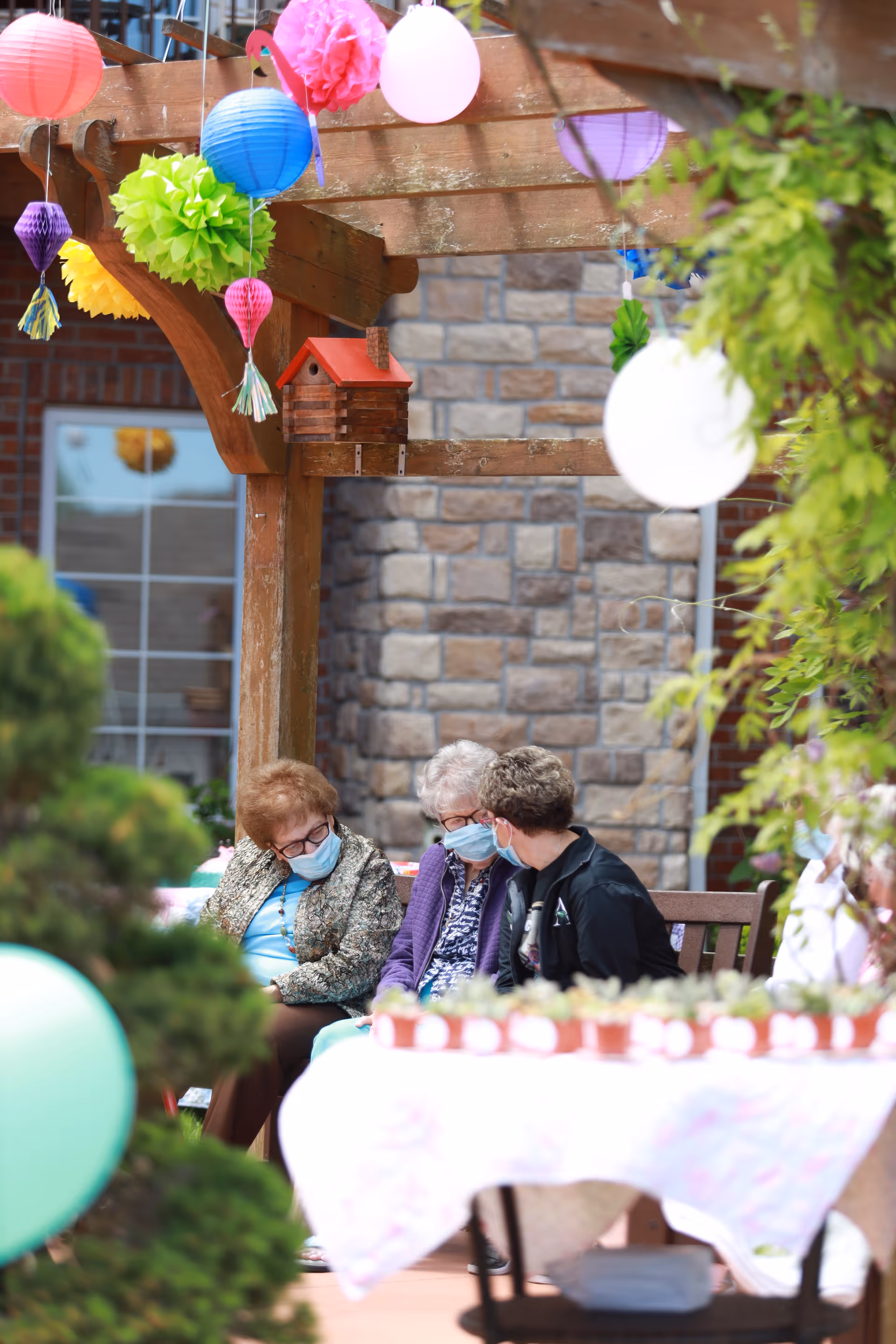 Three masked older adults sit and talk on a bench under a decorated wooden pergola with hanging paper lanterns and a table of potted plants.