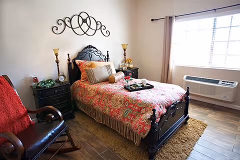A cozy bedroom with a dark wooden bed frame and a colorful patterned bedspread. The bed is adorned with multiple pillows and a tray with a cup and flowers. There are two matching nightstands with lamps and decorative items. A wooden rocking chair with a red throw blanket is positioned near the bed. A large window with curtains lets in natural light, and a wall-mounted air conditioning unit is below the window. The floor is wooden with a brown rug beside the bed.