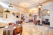 A spacious dining area in an assisted living facility with several round tables covered with brown tablecloths and set with napkins. The room features tiled floors, warm beige walls, decorative wall hangings including plants and utensils, and hanging light fixtures. There are arched doorways leading to other rooms and a welcoming atmosphere.