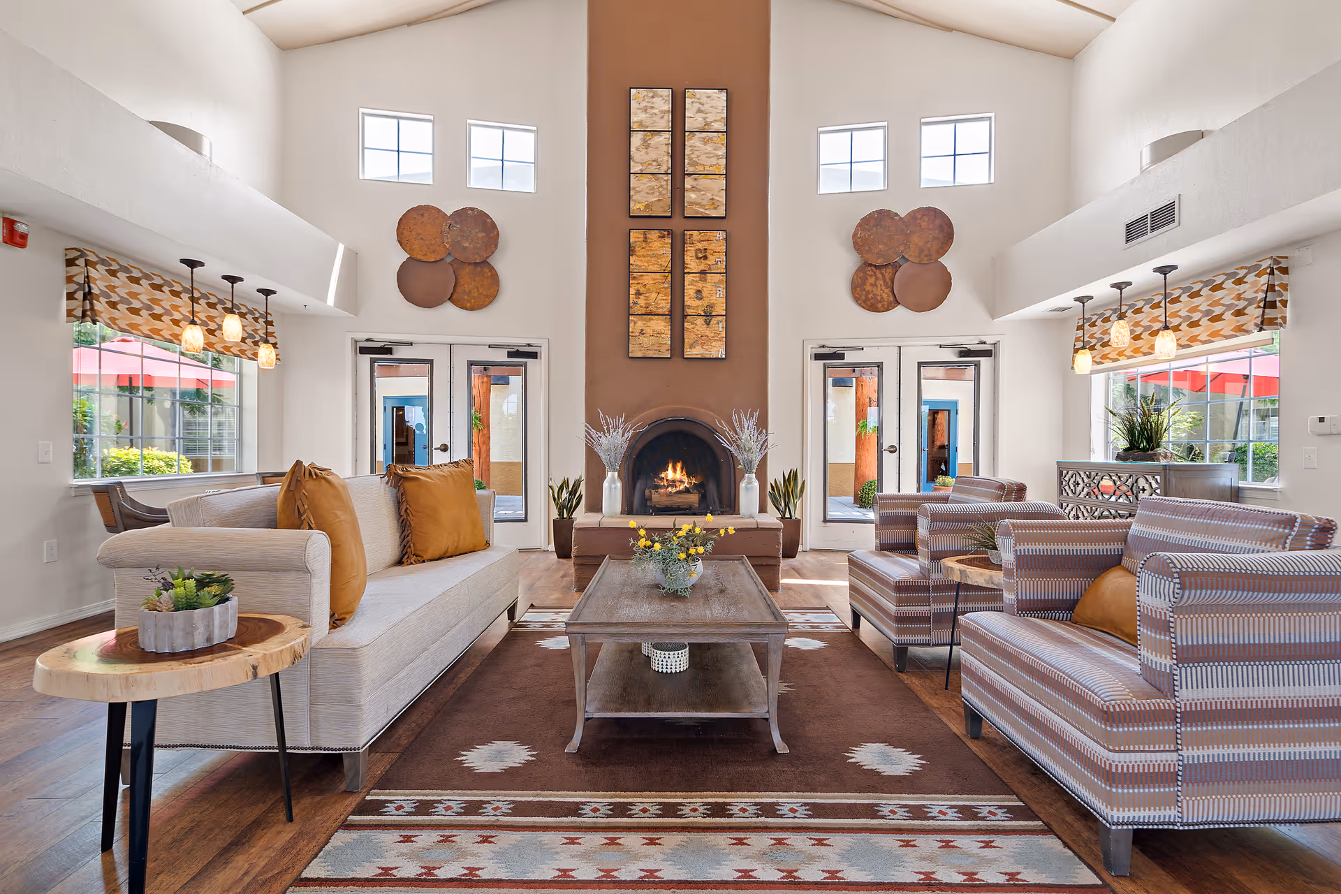 A bright and spacious living room with high ceilings and large windows. The room features a central fireplace with decorative art above it, two beige sofas with brown cushions, two striped armchairs, a wooden coffee table with a flower arrangement, and side tables with plants. The windows have patterned valances and pendant lights hanging above. Outside, red umbrellas and greenery are visible through the glass doors and windows.