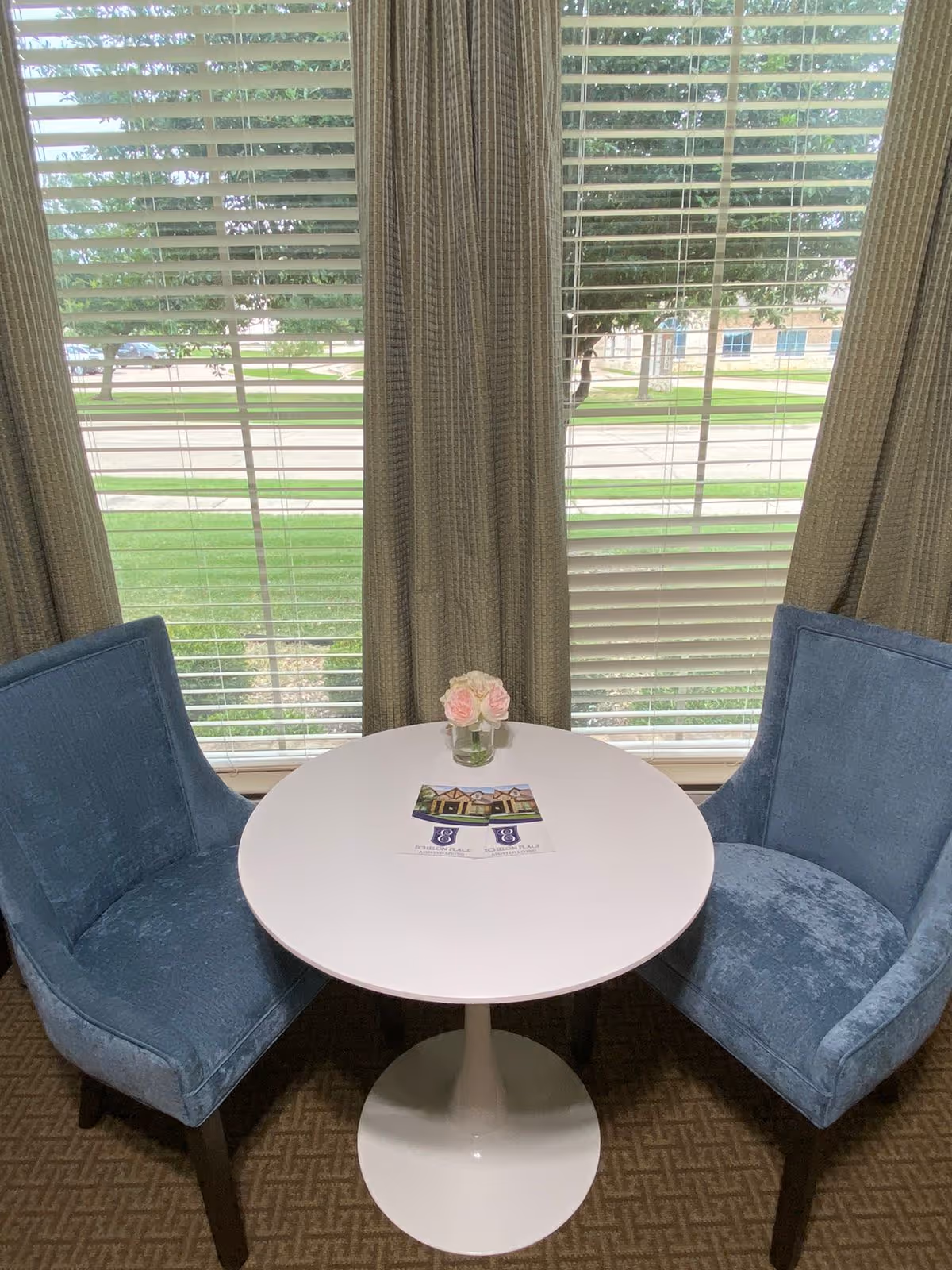 A small round white table with a vase of pink and white roses and two brochures on top, flanked by two blue upholstered chairs, positioned in front of a large window with beige curtains and white blinds, overlooking a green lawn and trees outside.